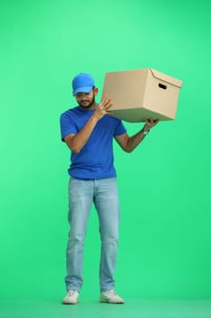 A male deliveryman, on a green background, in full height, shakes a box.