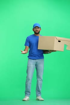 A male deliveryman, on a green background, in full height, with a box, throws up his hands.