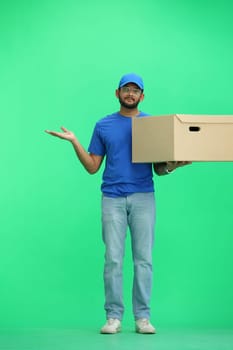 A male deliveryman, on a green background, full-length, with a box, points to the side.