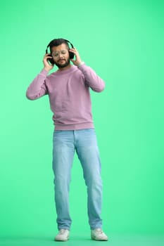 A man, on a green background, in full height, listening to music with headphones.