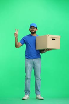 A male deliveryman, on a green background, in full height, with a box, shows a victory sign.