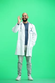 A male doctor, on a green background, in full height, shows the ok sign.