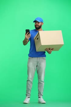 A male deliveryman, on a green background, in full height, with a box and a phone.