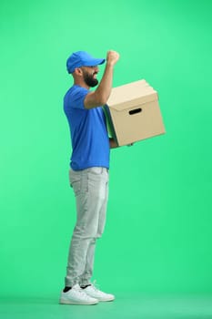 A male deliveryman, on a green background, full-length, with a box, raised his hand up.