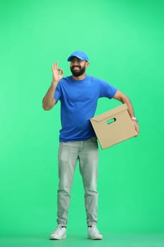 A male deliveryman, on a green background, in full height, with a box, shows the ok sign.