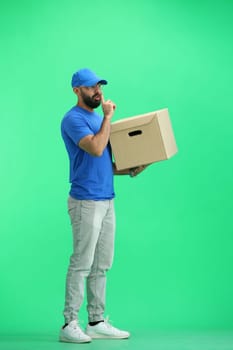 A male deliveryman, on a green background, in full height, with a box, shows a sign of silence.