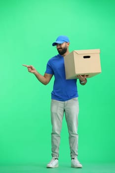 A male deliveryman, on a green background, full-length, with a box, points to the side.