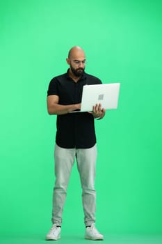 A man, on a green background, in full height, uses a laptop.