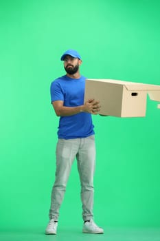 A male deliveryman, on a green background, in full height, shakes a box.