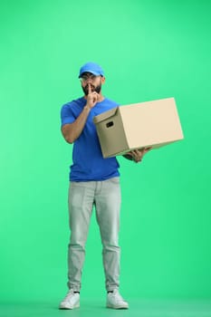 A male deliveryman, on a green background, in full height, with a box, shows a sign of silence.
