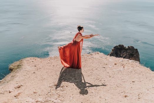 Woman red dress sea. Female dancer posing on a rocky outcrop high above the sea. Girl on the nature on blue sky background. Fashion photo