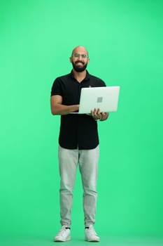 A man, on a green background, in full height, uses a laptop.