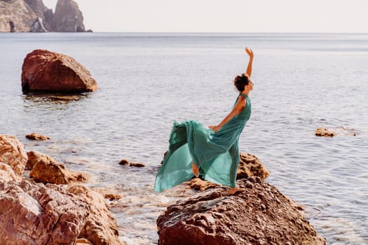 Woman green dress sea. Woman in a long mint dress posing on a beach with rocks on sunny day. Girl on the nature on blue sky background