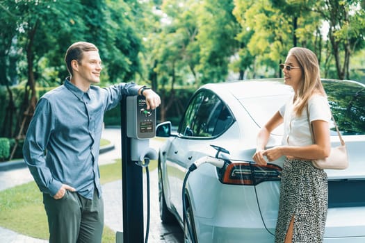 Young couple travel with EV electric car charging in green sustainable city outdoor garden in summer shows urban sustainability lifestyle by green clean rechargeable energy of electric vehicle innards