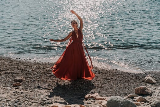 Woman red dress sea. Female dancer in a long red dress posing on a beach with rocks on sunny day. Girl on the nature on blue sky background