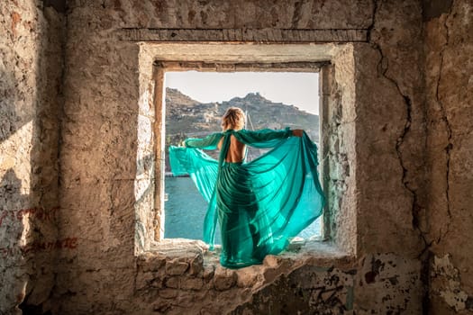 Rear view of a happy blonde woman in a long mint dress posing against the backdrop of the sea in an old building with columns. Girl in nature against the blue sky