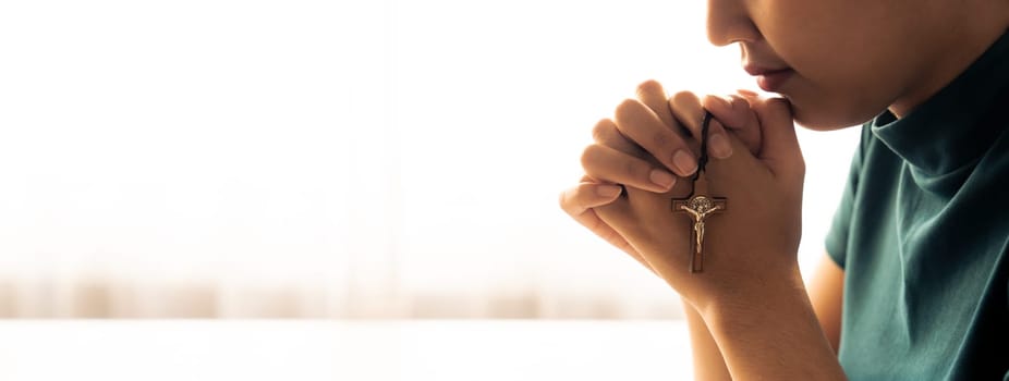 Praying female hand holding cross while praying to god faithfully. Concept of hope, religion, faith, christianity and god blessing for happiness. Bright light, white blurring background. Burgeoning.