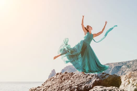 Side view a Young beautiful sensual woman in a red long dress posing on a rock high above the sea during sunrise. Girl on the nature on blue sky background. Fashion photo.