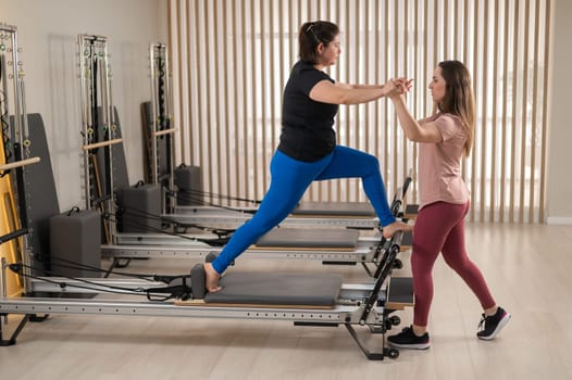 Overweight caucasian woman doing pilates exercises on reformer with personal trainer