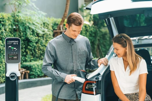 Young couple travel with EV electric car charging in green sustainable city outdoor garden in summer shows urban sustainability lifestyle by green clean rechargeable energy of electric vehicle innards