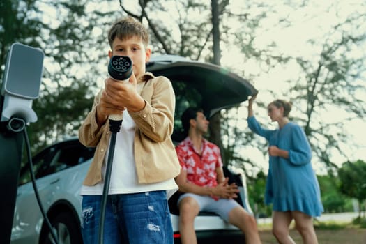 Little boy holding EV charger and point at camera with his family sitting on the trunk in background. Road trip travel with alternative energy charging station for eco-friendly car concept. Perpetual