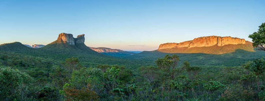 A stunning panoramic view of the typical landscape of Chapada Diamantina, with rocky hills shaped like camels, lush forests, and valleys at sunset, with a clear blue sky providing ample space for text.