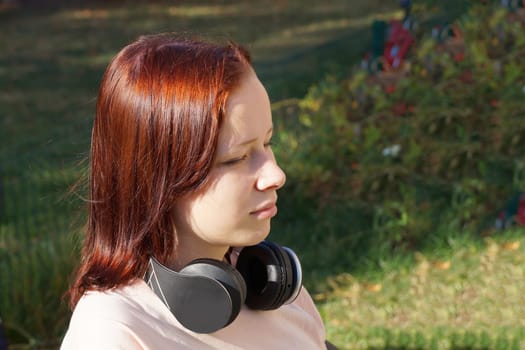red-haired teenage girl sitting with her eyes closed in headphones in the park.