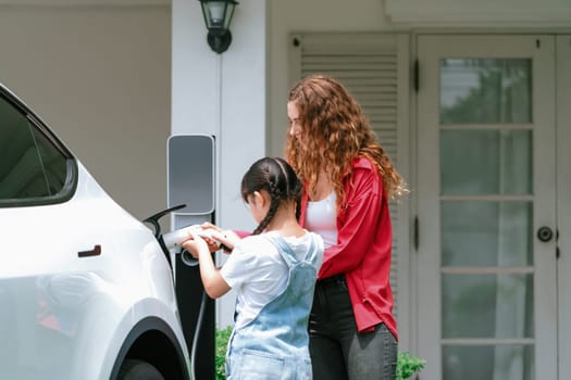 Happy little young girl learn about eco-friendly and energy sustainability as she help her mother recharge electric vehicle from home EV charging station. EV car and modern family concept. Synchronos