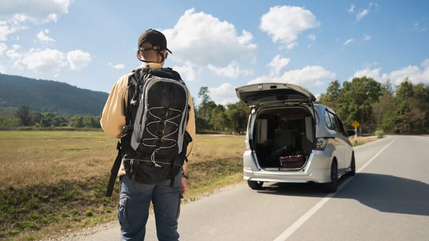 Rearview shot of male traveler with backpack walking towards a car at countryside with blue sky.