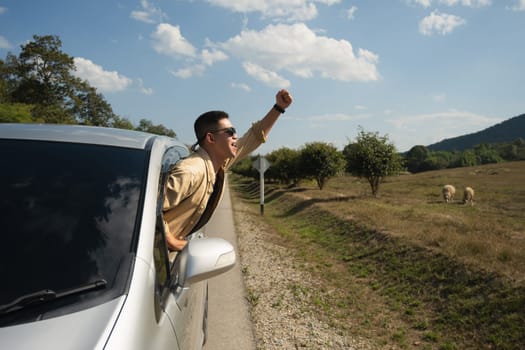 Happy Asian man in sunglasses leaning out from car window against blue sky. Travel and transportation concept.