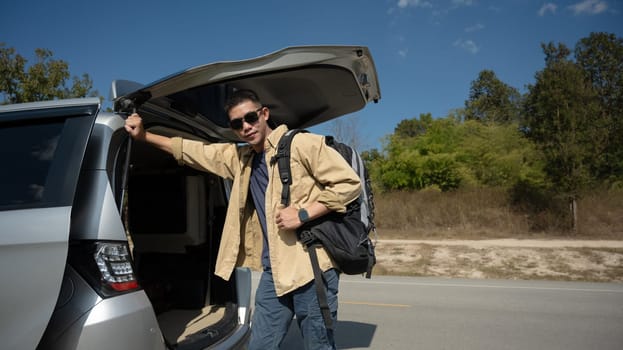 Portrait of smiling Asian man in sunglasses standing near open trunk of car. Travel and transportation concept.