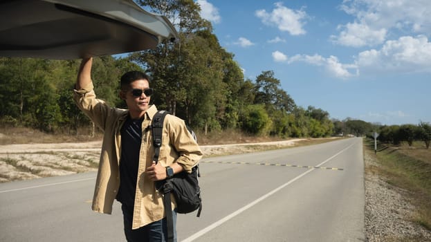 Smiling young man with backpack opening a trunk door of his car at countryside with blue sky.