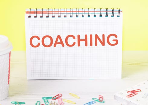 COACHING the word is written on a blank sheet in a notebook standing on a table on a yellow background