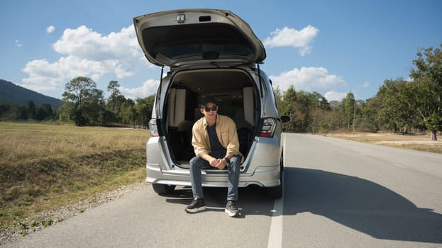 Handsome young man sitting on the open trunk of his car against blue sky and white cloud.