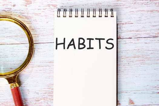 HABITS a word written on a notebook on wooden boards next to a magnifying glass