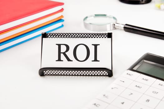 Return on investment concept. ROI text on a business card next to office supplies on a white background