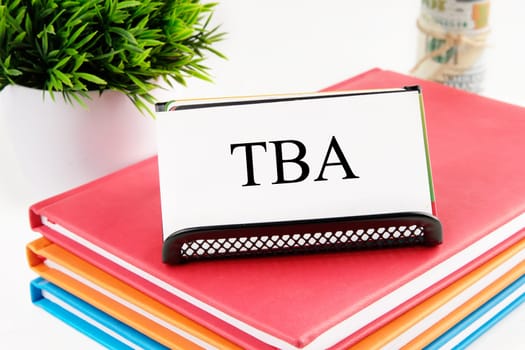 TBA text on a white business card standing on a stand on notebooks, next to the money out of focus and a green plant on a white background
