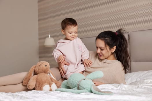 happy mother and her little child daughter playing with bunny toys in bedroom, family having fun