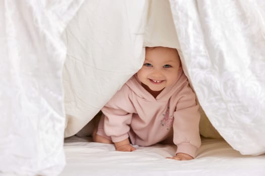 cute funny little child girl looks out from under the blanket on the bed, copy space