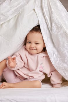 cute funny little child girl looks out from under the blanket on the bed