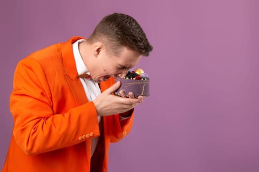 handsome birthday man in orange jacket eating cake on purple background. copy space