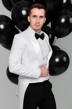 elegant caucasian man in white suit tuxedo with bunch of black air balloons on white background. holiday celebration