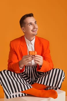 extravagant funny young man in orange jacket and striped pants holding cupcake on orange background.