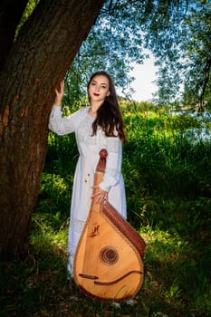 A young bandurist in a white dress. A female bandurist with a bandura is standing near a tree on the bank of the river