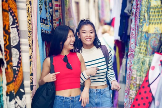 Smiling young women friends in casual clothes walking along colorful clothing store on street market in city during summer vacation