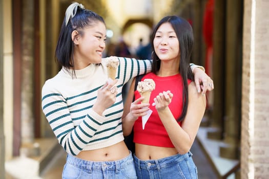Positive Asian woman standing with hand on shoulder of friend while eating delicious ice cream in street against blurred background
