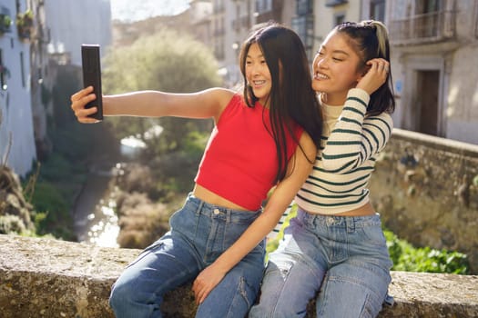 Positive young Asian women taking self shot on cellphone while having fun together and sitting near building in Granada