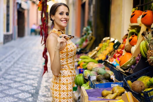 Side view of positive young female greengrocer, in casual yellow gown and makeup standing near stall with fresh fruits on paved street at town in daylight