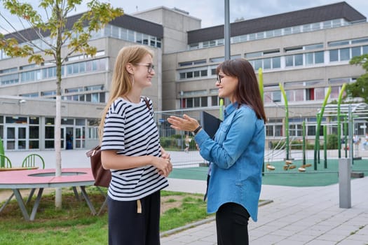 Talking female teacher and teenage high schoolgirl outdoor, school building background. Meeting communication student girl with backpack and mentor counselor. Education, adolescence, learning concept