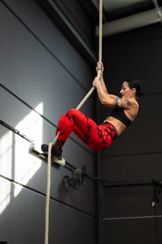Vertical photo of a strong mature woman with tattooed arms climbing a rope in a cross training gym